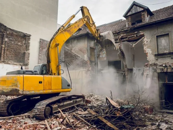  A yellow excavator demolishing an old, brick building, creating a pile of rubble and dust.