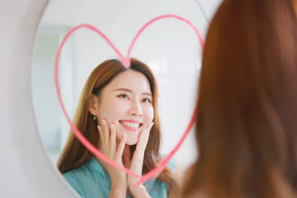 Smiling woman looking at her reflection in the mirror framed by a heart, symbolizing self-love, body confidence, and intimacy empowerment.