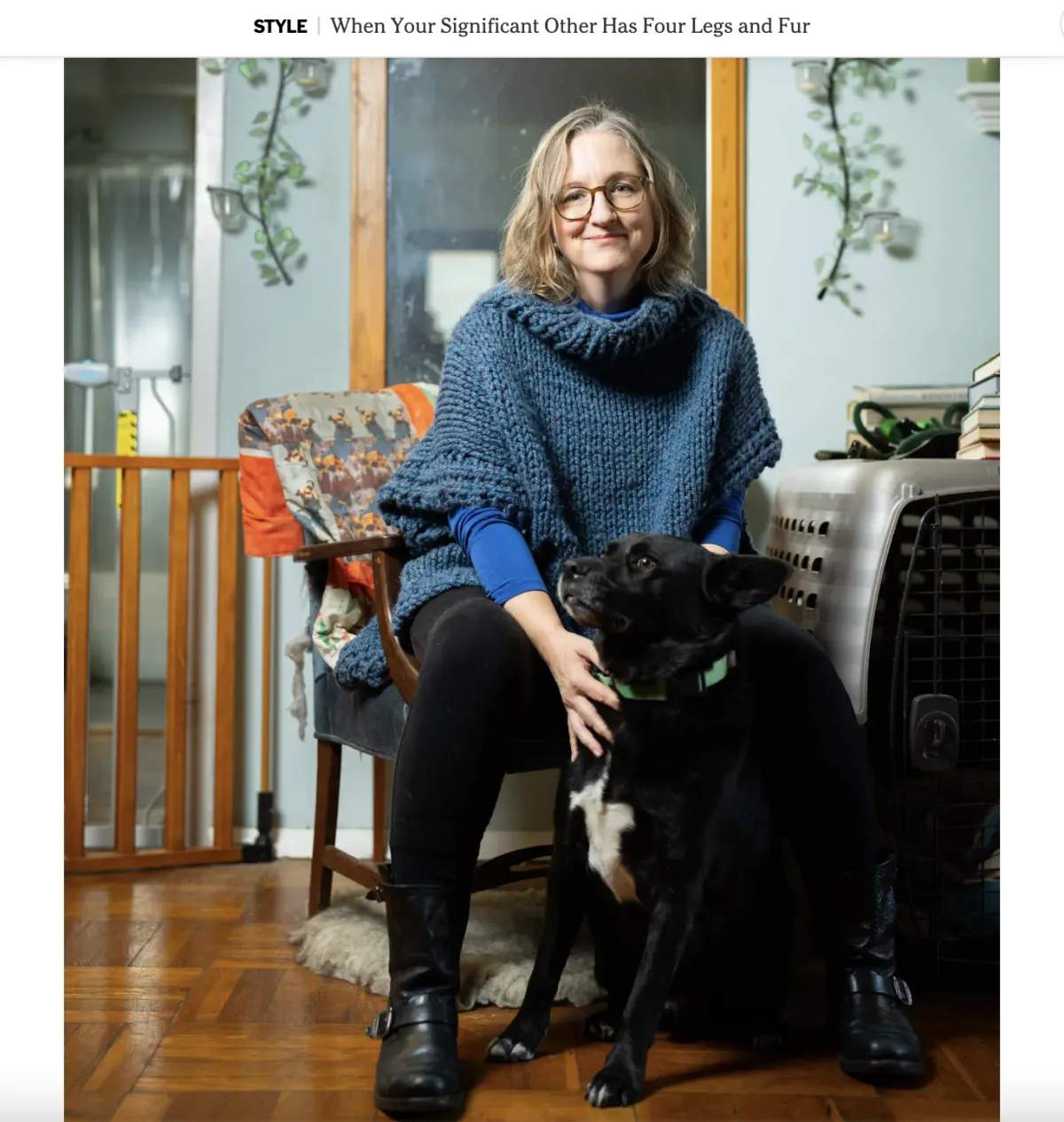 A woman in a blue sweater and black leggings sits in a chair with a black dog sitting on the floor in front of her.