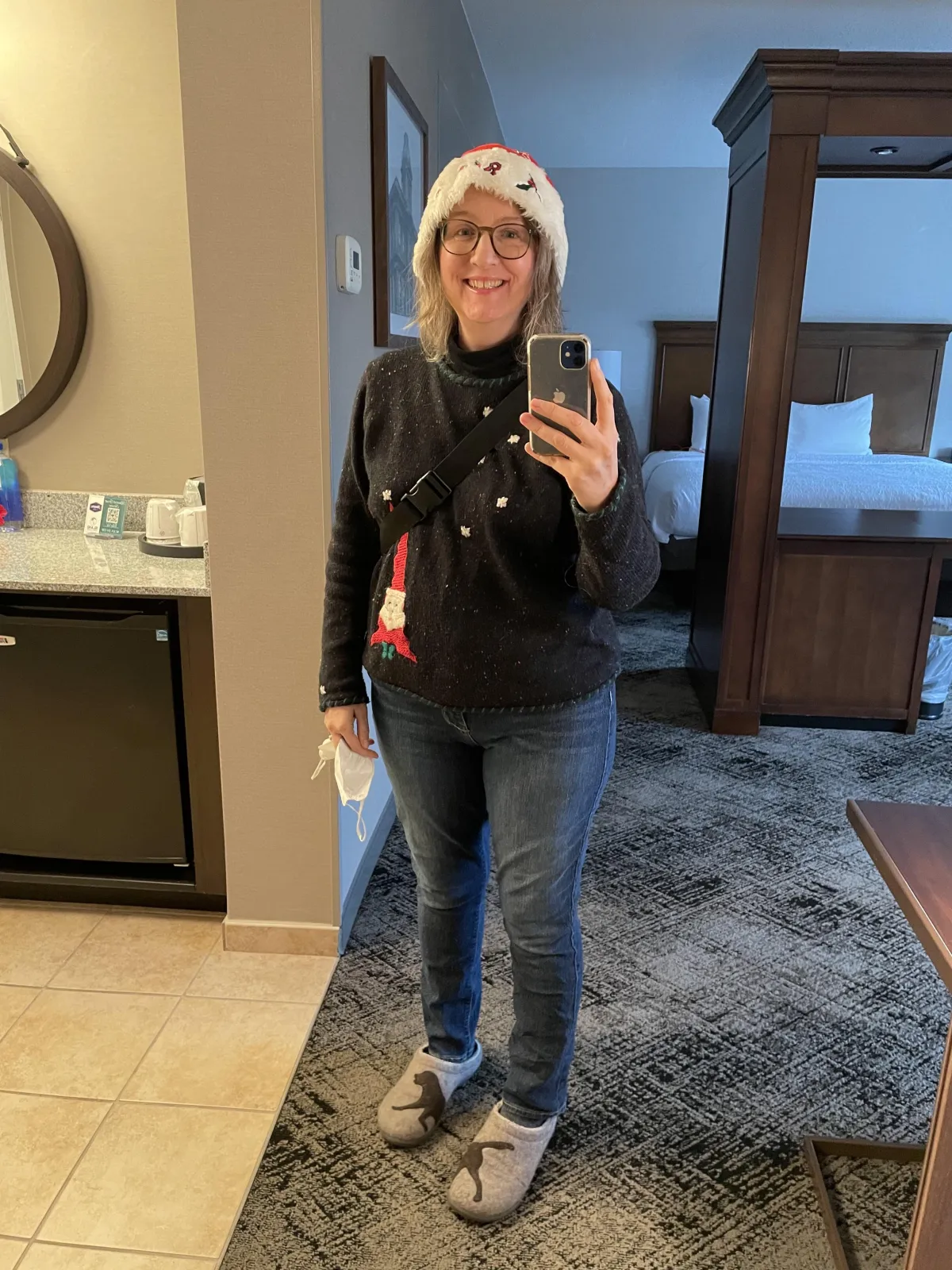 Kizz stands in a hotel room wearing Jeans, a Christmas sweater, and a Santa Hat.