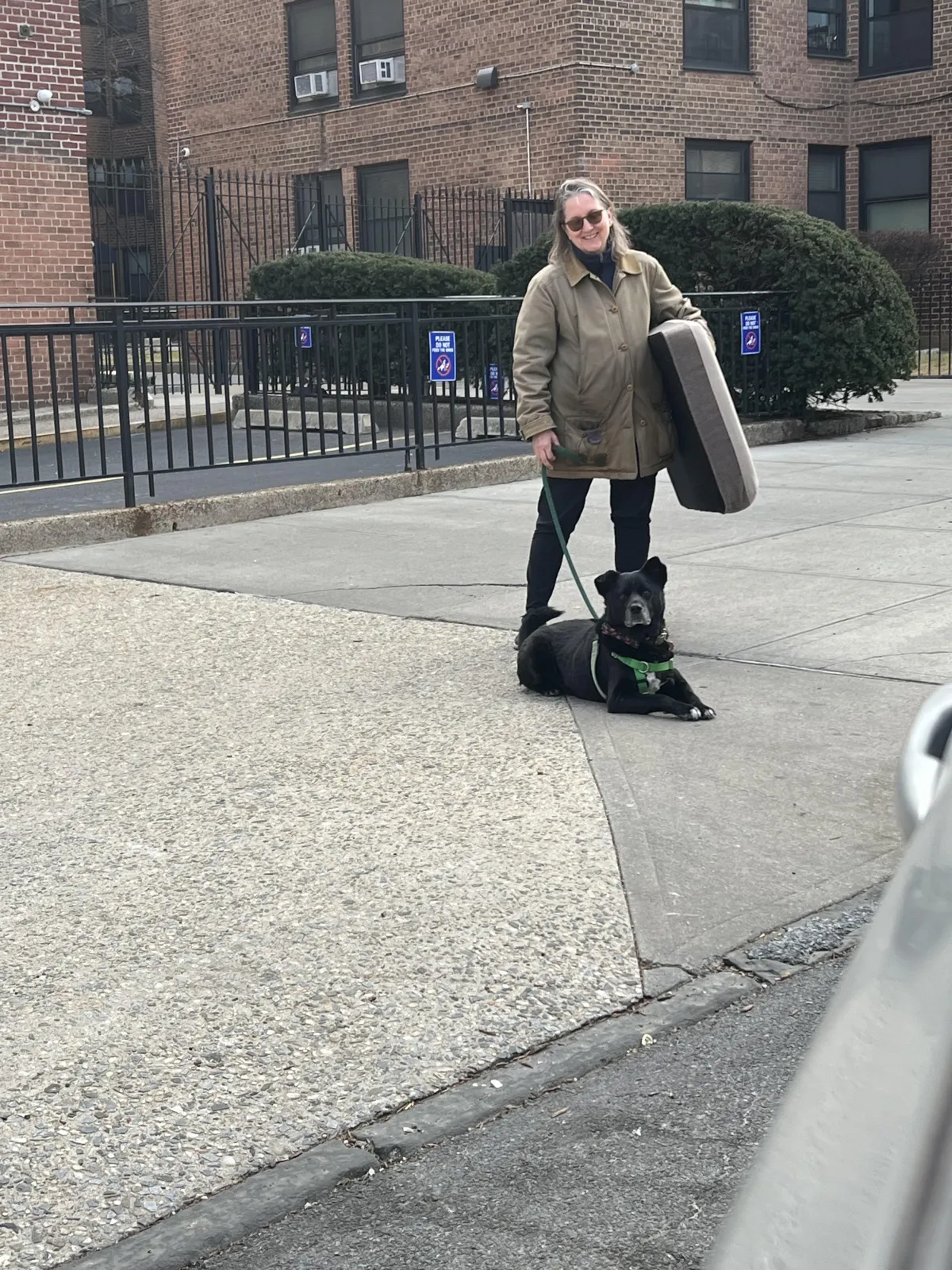 A woman in black pants, a green barn coat & dark glasses smiles. She holds a dog bed under one arm and a leash in the other hand. A fuzzy black medium-sized dog is on the leash. He is lying down on the sidewalk.