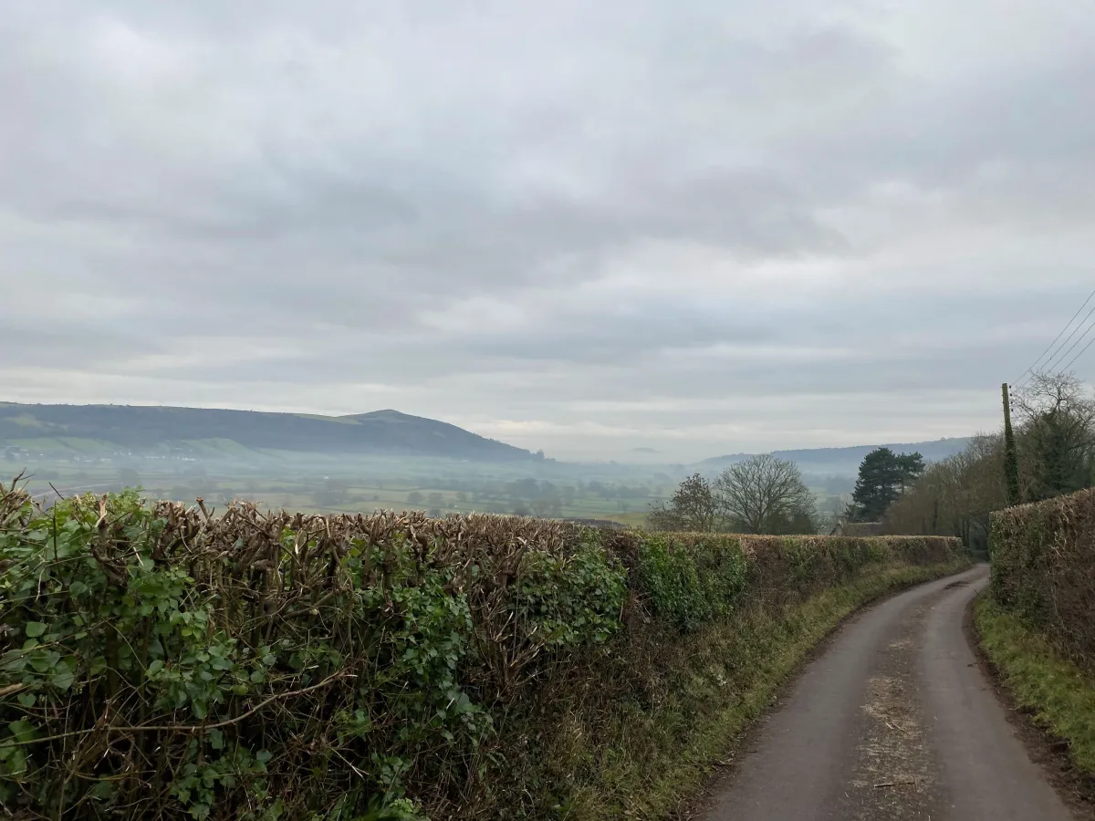 A peaceful winter countryside scene with a misty hillside, quiet country lane, and expansive grey, cloudy view across the landscape.