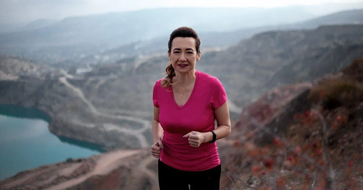 An older woman in workout clothing walks up a trail. There is a body of water and some mountains behind her.