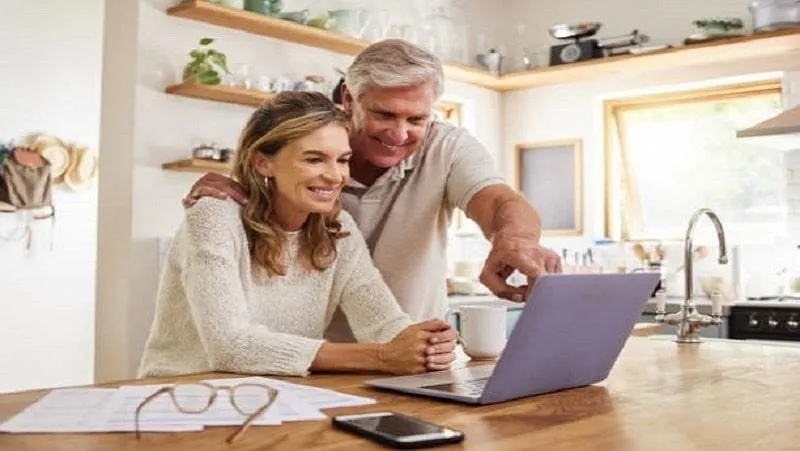 Pareja de mediana edad revisando beneficios fiscales del seguro de vida en una computadora portátil en su cocina. Están felices y sonrientes, con documentos y un teléfono móvil en la mesa, en un ambiente luminoso y moderno.