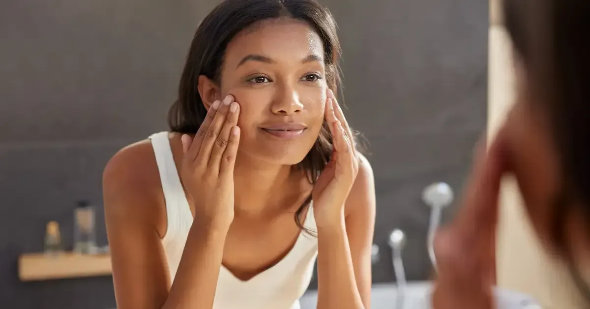 Woman with sensitive skin gently applying moisturizer to her face in front of a bathroom mirror