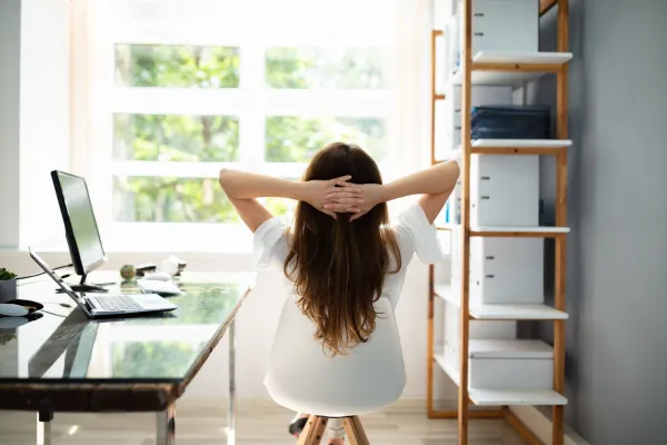 A female bookkeeper with long hair sitting at her home office desk, relaxing with her hands clasped behind her head, looking out a window beside a shelf with binders and a laptop open next to her.