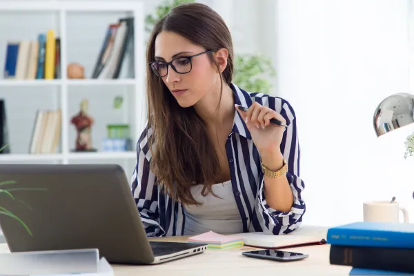 A focused female virtual bookkeeper with glasses, wearing a striped blazer, working intently at her laptop in a well-organized home office.