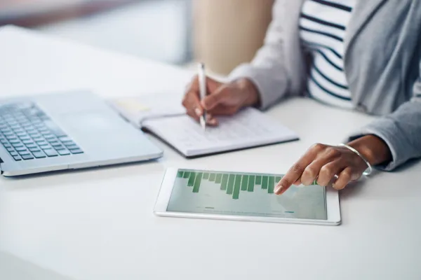 A virtual bookkeeper in a gray blazer using a tablet to analyze a bar graph while taking notes in a notebook, with a laptop beside them on the desk.