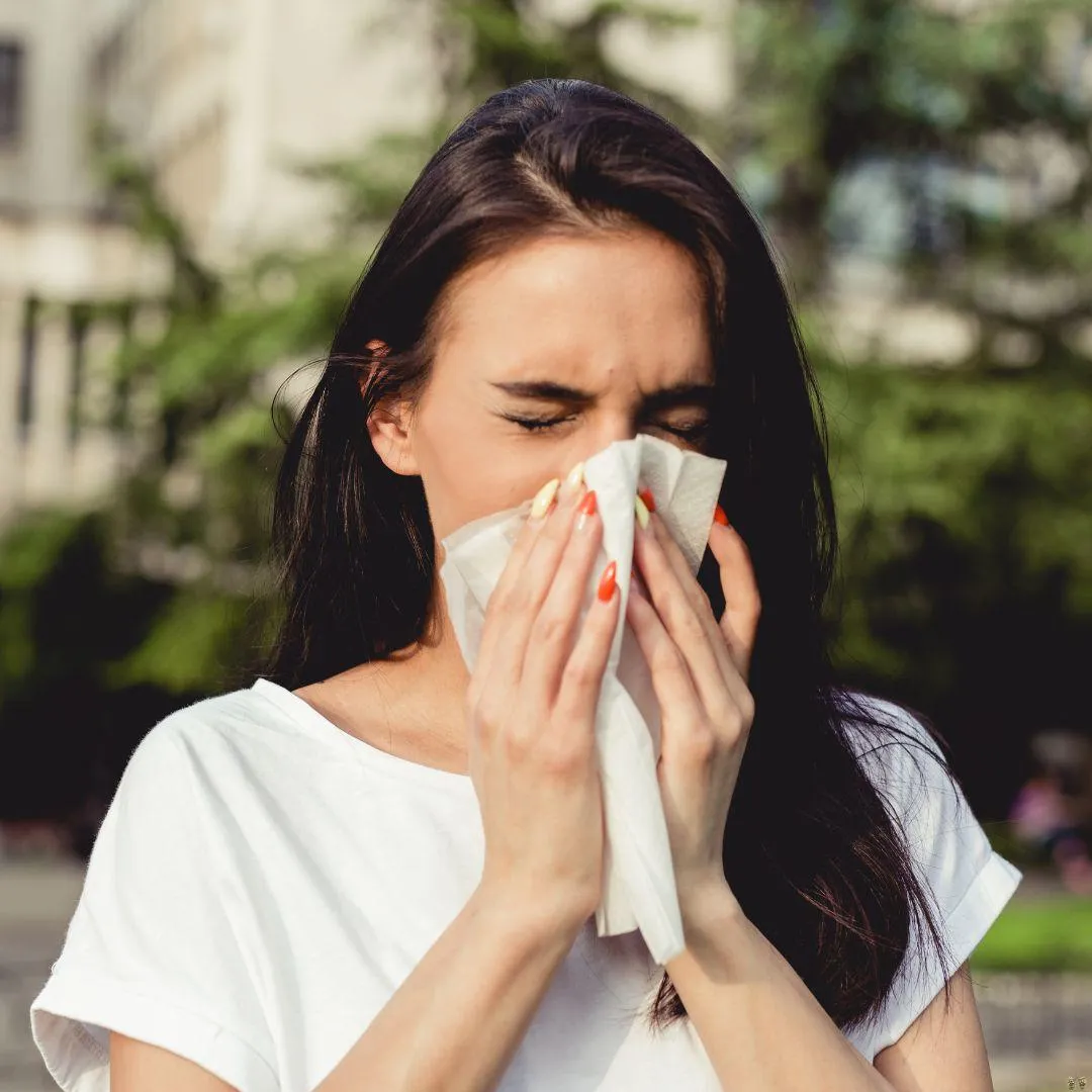A young woman with long dark hair is sneezing into a tissue while standing outdoors. She is wearing a white T-shirt and appears to be experiencing allergy symptoms. Trees and a building are visible in the blurred background.
