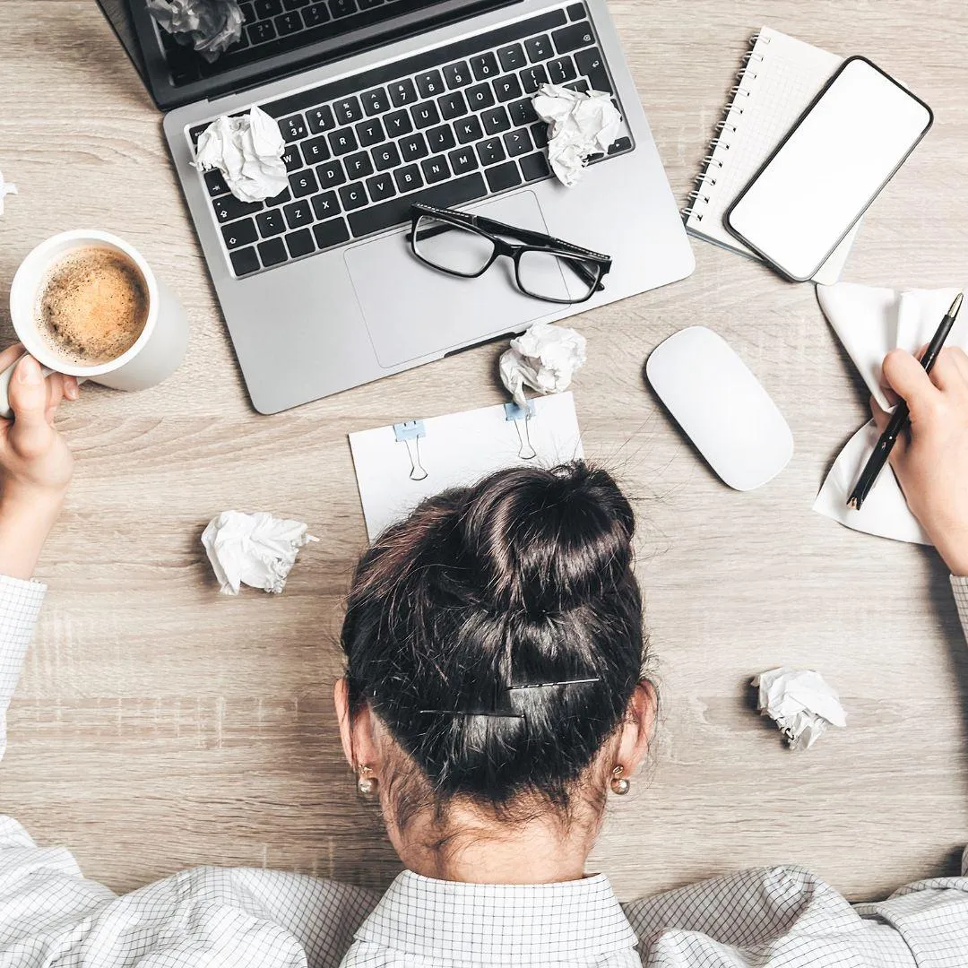 A woman with dark hair in a bun sits at a cluttered desk, appearing overwhelmed. Her head is down, resting on the desk, surrounded by crumpled papers, a laptop, a cup of coffee, glasses, a notebook, a smartphone, a computer mouse, and a pen in hand.