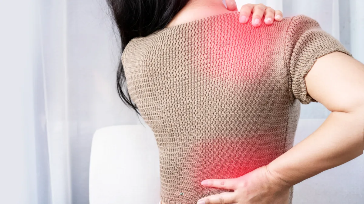 A woman with long dark hair is holding her upper back and lower back with both hands, indicating pain. Red highlights on the shoulder and lower back suggest areas of discomfort or inflammation.