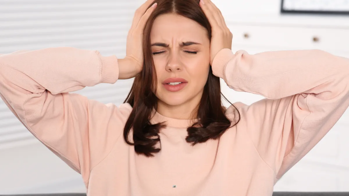 A woman wearing a light pink sweatshirt is holding her head with both hands and has her eyes closed, expressing discomfort or pain, possibly due to a headache or stress.