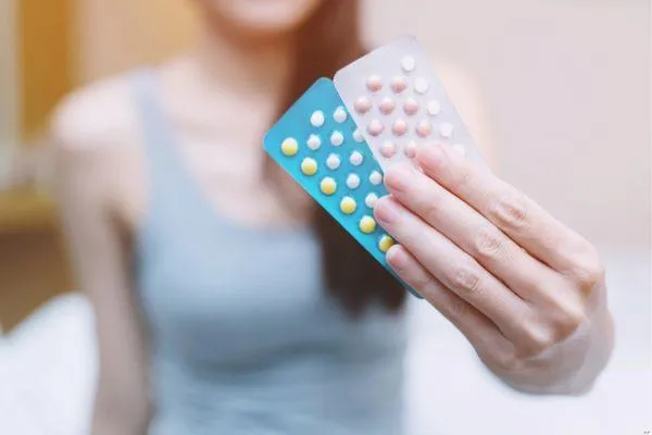Woman holding two blister packs of birth control pills in different colors, wearing a gray tank top, with the focus on the hand and pills and the background softly blurred.