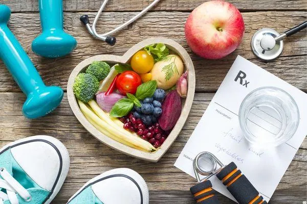 Flat lay of healthy lifestyle essentials including a heart-shaped bowl filled with fruits and vegetables, a stethoscope, dumbbells, a jump rope, sneakers, an apple, a glass of water, and a prescription pad suggesting fruits and vegetables.