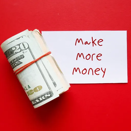 A red background with a white note card that reads: "Make More Money." A roll of money is sitting next to the note card.