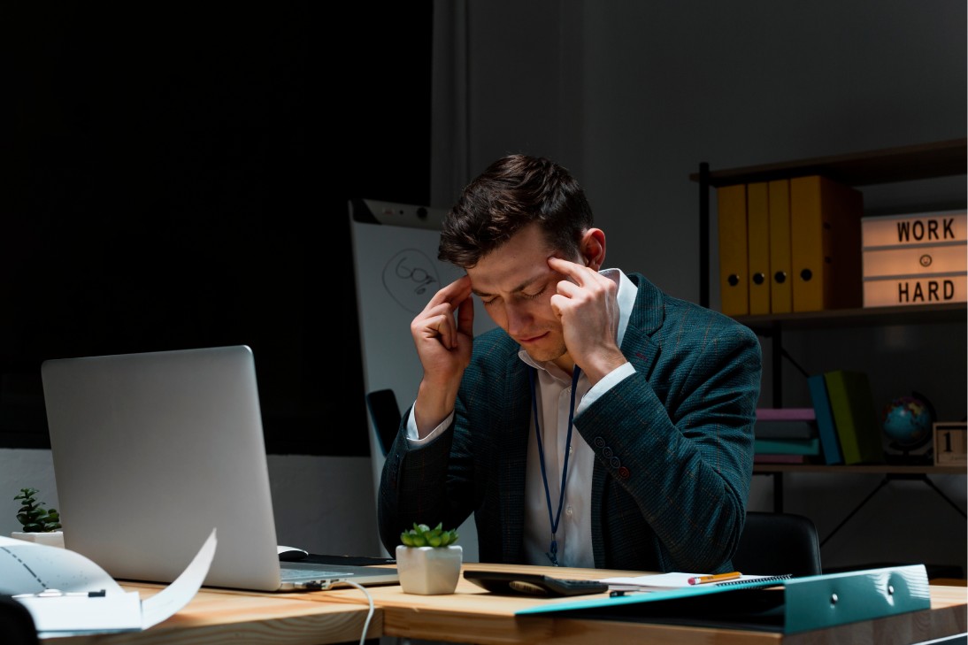 Man in office, wearing a suit, sits at a wooden desk, rubbing his temples in frustration. A laptop is open, and a Work Hard sign is visible.