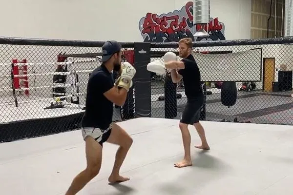 Two fighters training in a caged MMA ring, one holding focus mitts while the other practices strikes, with a modern gym setup in the background showcasing equipment and motivational artwork