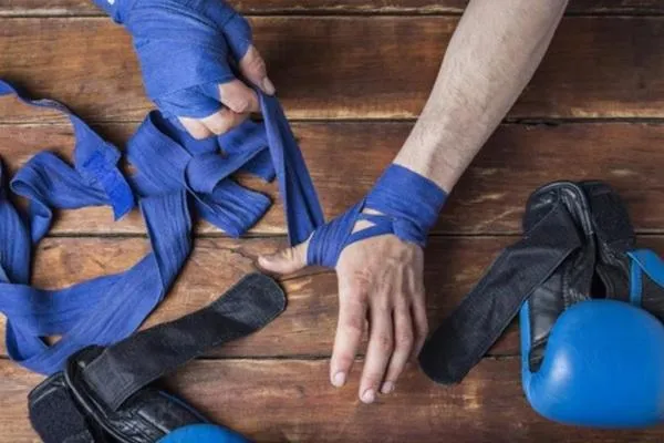 A fighter wrapping their hands with blue hand wraps on a wooden surface, with protective MMA gloves beside them, symbolizing injury prevention and proper preparation.