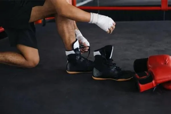 A fighter sitting on the edge of a training mat, wrapping their hands in preparation for a fight. Nearby are black boxing shoes being tied and red boxing gloves resting on the ground, symbolizing readiness for combat.