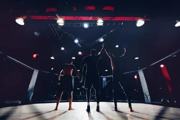 An MMA referee raises the hand of a victorious fighter in the cage, symbolizing the journey from amateur to professional competition under bright arena lights.