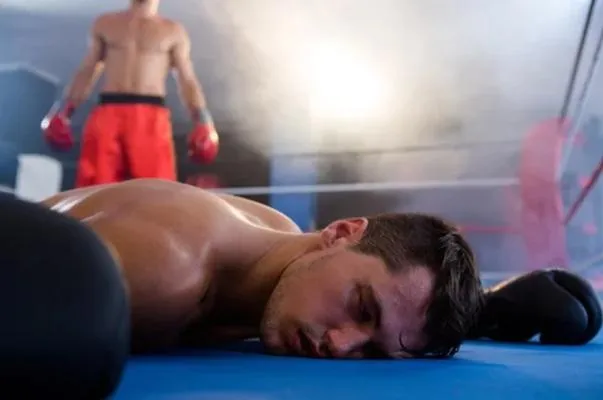 A knocked-out MMA fighter lying face-down on the canvas, with an opponent standing in the background, symbolizing the risks of concussions in combat sports.