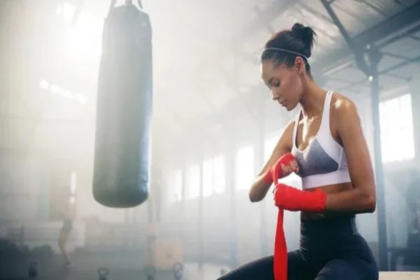 Female fighter wrapping her hands in red wraps inside a sunlit gym, preparing for intense training or a fight.