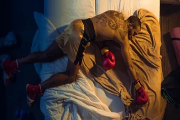 MMA fighter wearing boxing gloves and shorts sleeping face-down on a bed after an intense training session, symbolizing the importance of recovery.