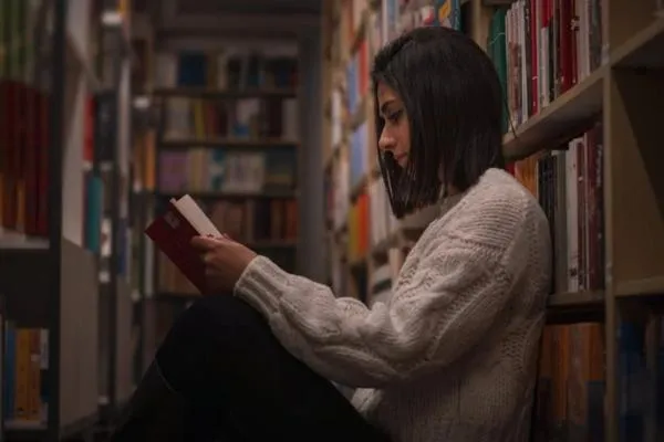 Young woman reading a book in a cozy library setting, surrounded by shelves filled with colorful books, illustrating the importance of reading and educational exploration for young learners.