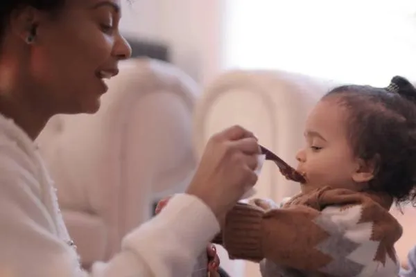 Mother feeding toddler with a spoon in a cozy indoor setting, emphasizing responsive feeding practices for young children.