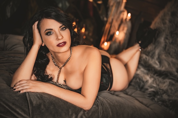 Woman posing confidently on a bed during a boudoir photography session, wearing black lingerie in a softly lit studio.