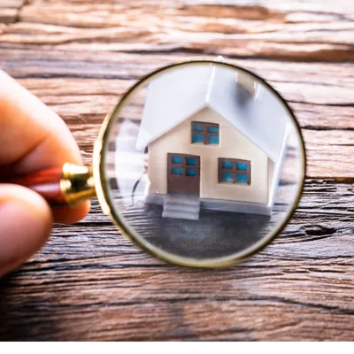 Close-up of a hand holding a magnifying glass over a miniature house on a wooden surface, symbolizing home value assessment or property analysis.