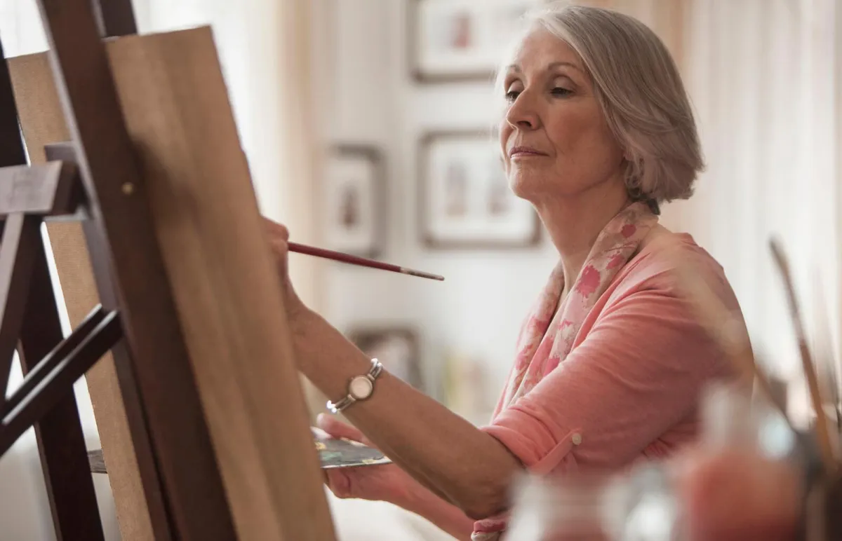 An elderly woman with silver hair, wearing a pink blouse and a floral scarf, painting on a canvas in a well-lit hobby room. She holds a paintbrush with a focused expression, surrounded by art supplies, brushes, and framed pictures, creating a warm and inspiring creative space