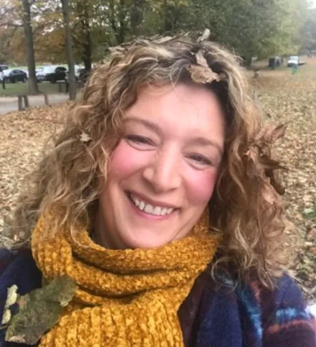 Rachel smiling at the camera wearing a mustard coloured scarf and autumnal coat among trees and leaves