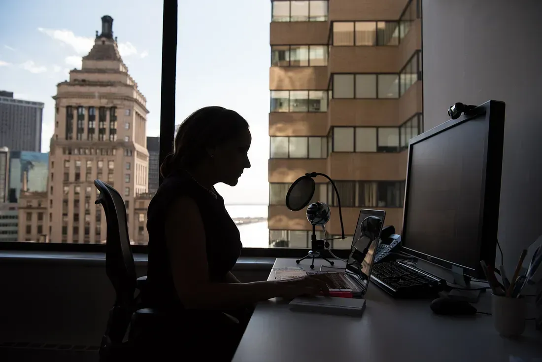 A woman sits at a desk in a high-rise office, working on her laptop with a microphone nearby, silhouetted against a large window overlooking the city.