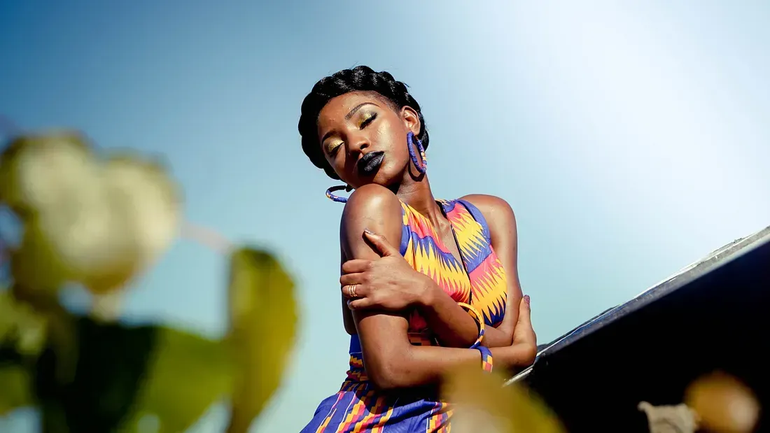 A Black woman in a colorful African print outfit poses with her arms crossed, eyes closed, and head tilted under a bright sky.