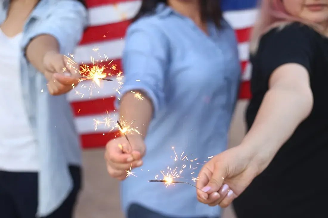 Three people holding sparklers in front of an American flag, celebrating with bright sparks in their hands.