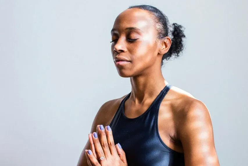 A woman in a black tank top practices mindfulness with her eyes closed and hands in a prayer position, exuding peace and relaxation.