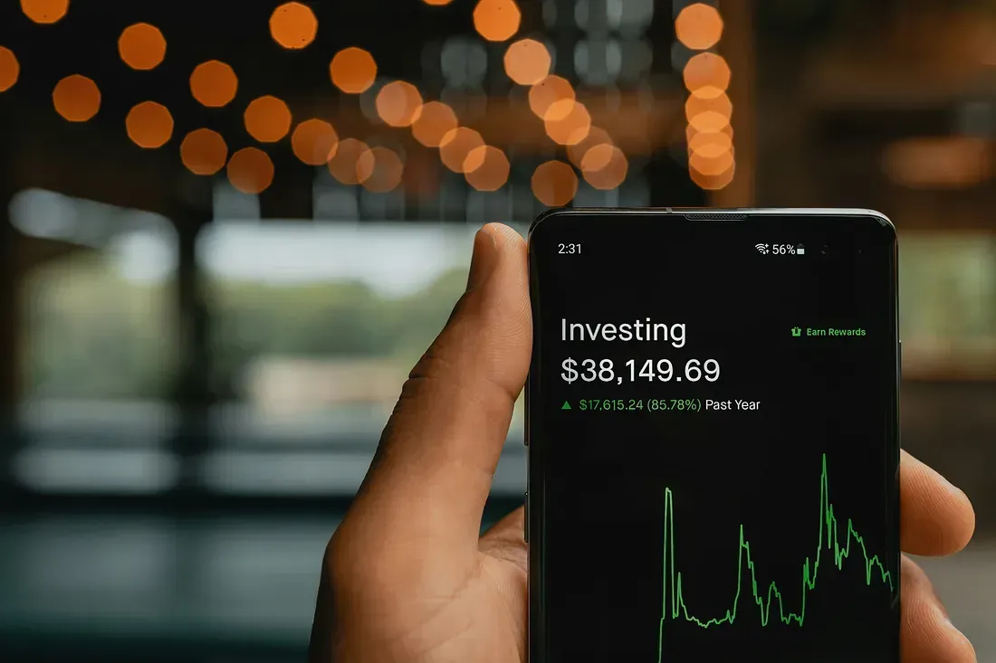 A hand holding a smartphone displaying an investment portfolio with a balance of $38,149.69 and an 85.78% gain over the past year.