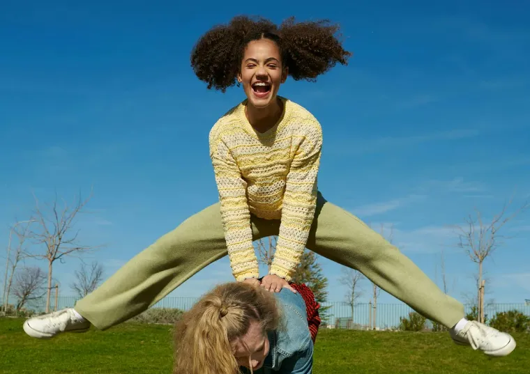 Two young women playing leapfrog outdoors on a sunny day, one jumping with a big smile while the other bends down.