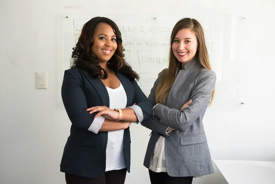 Two professional women standing confidently with arms crossed in front of a whiteboard, smiling at the camera in a business setting.