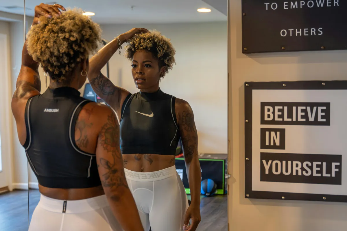 A Black woman with short curly hair looks at herself in the mirror at a gym, standing near a "Believe in Yourself" sign.