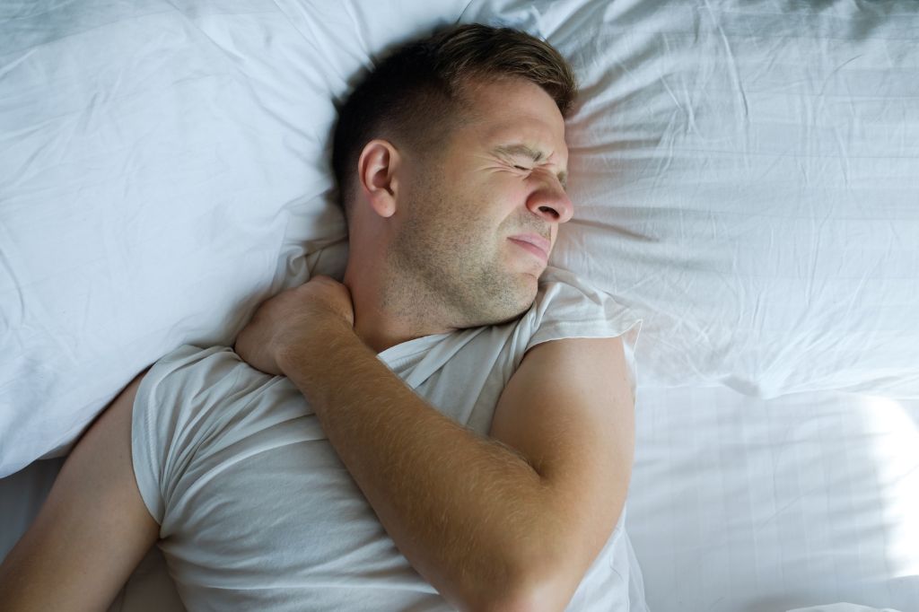 Person lying awake in bed holding their shoulder in discomfort, showing shoulder pain that worsens at night while trying to sleep
