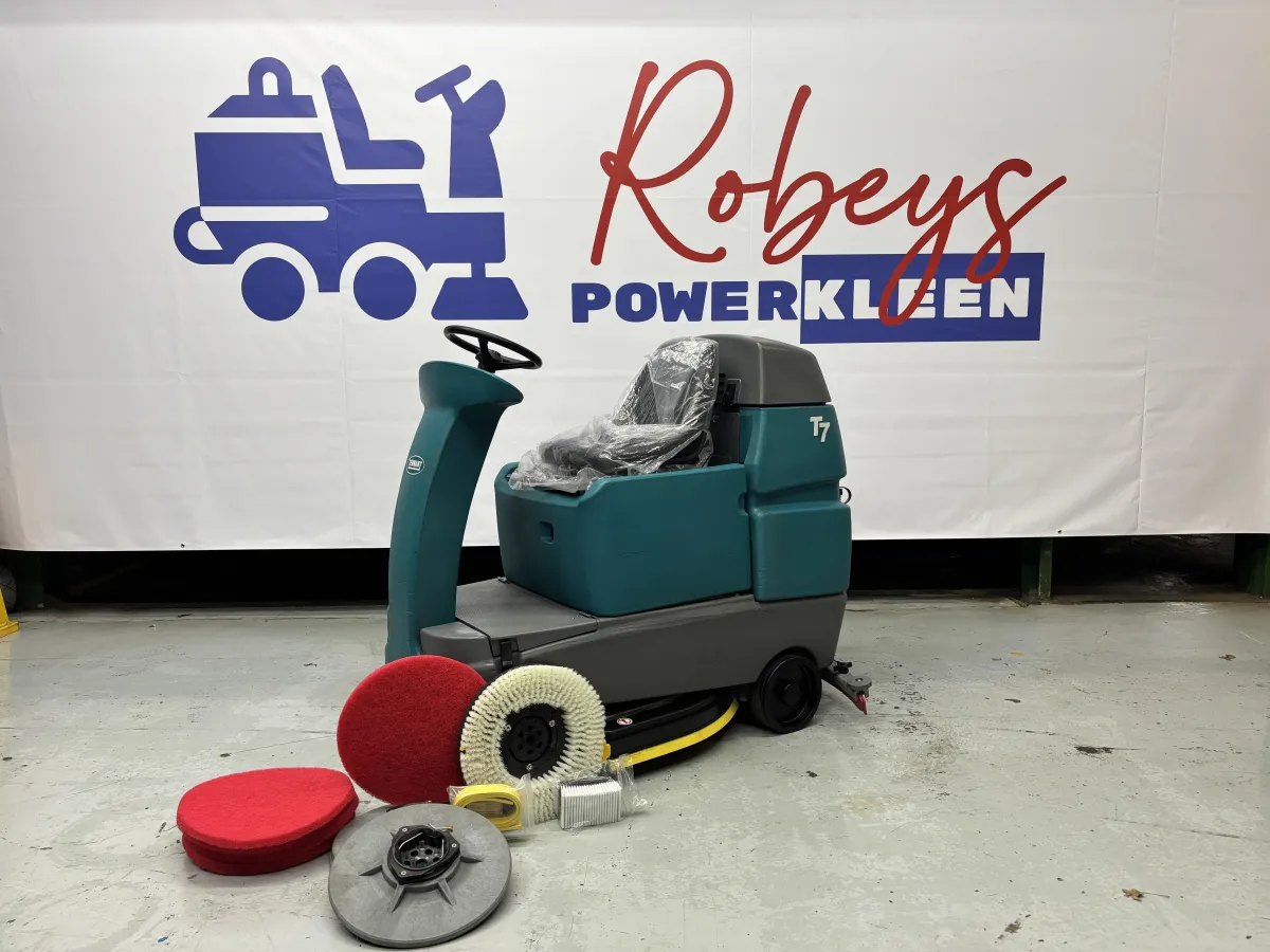 Tennant T7 ride-on floor scrubber positioned on a warehouse floor, surrounded by red scrubbing pads, a white nylon bristle brush, and a drive disk.