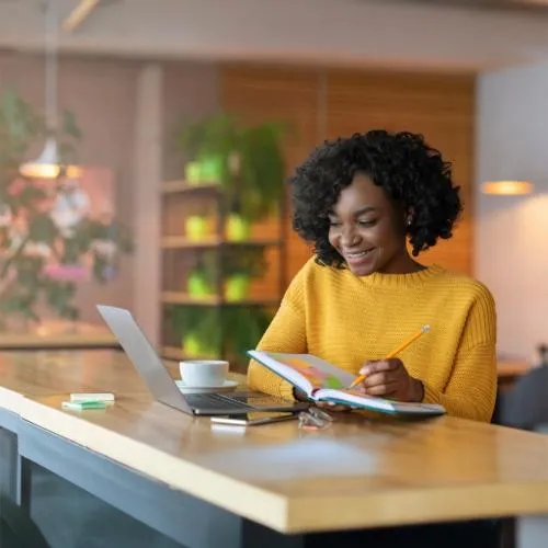 A young professional smiling while working on her laptop and notebook in a cozy, modern workspace with plants and warm lighting in the background, representing productivity and job preparation.