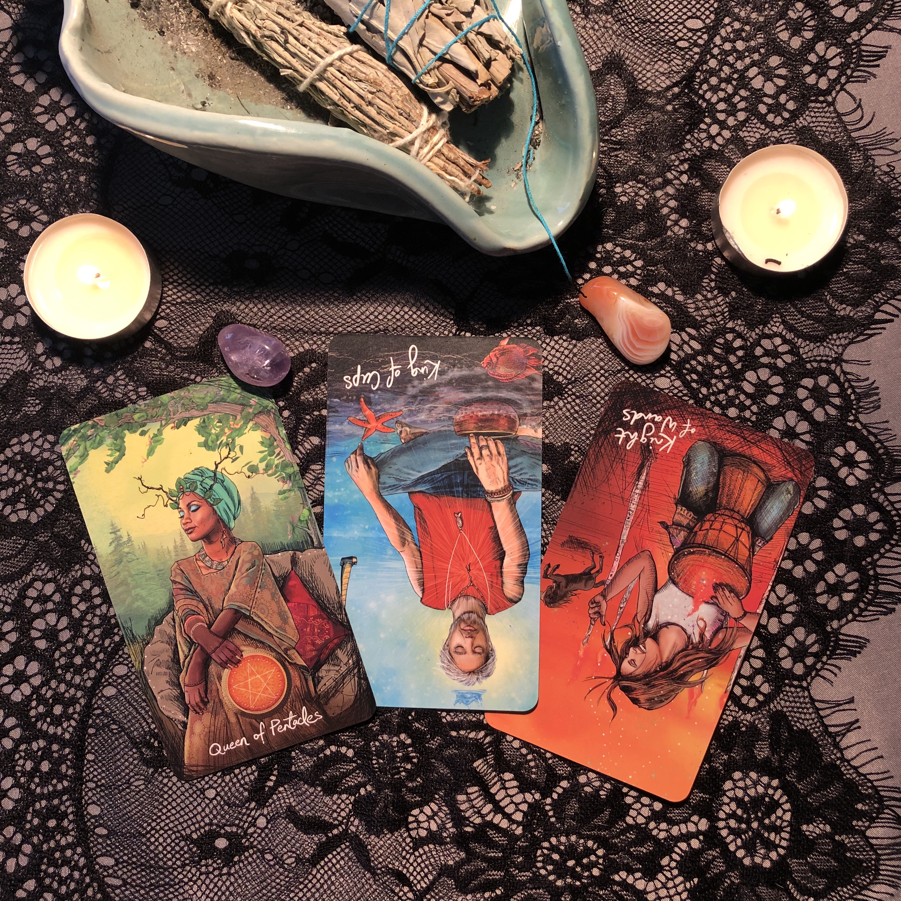 Three tarot cards from The Light Seer's Tarot spread on black lace with crystals, candles, and palo santo - Queen of Pentacles, reversed King of Cups, and reversed Knight of Wands