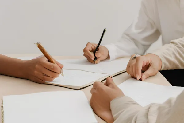 Three individuals engaged in a collaborative discussion, writing notes in open notebooks on a table. Their hands and pens are visible, suggesting teamwork, planning, or a professional meeting. Select 72 more words to run Humaniser.