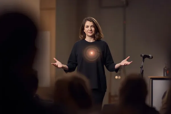 a confident speaker standing on a small stage and in front of a microphone, speaking to an audience with calm expression