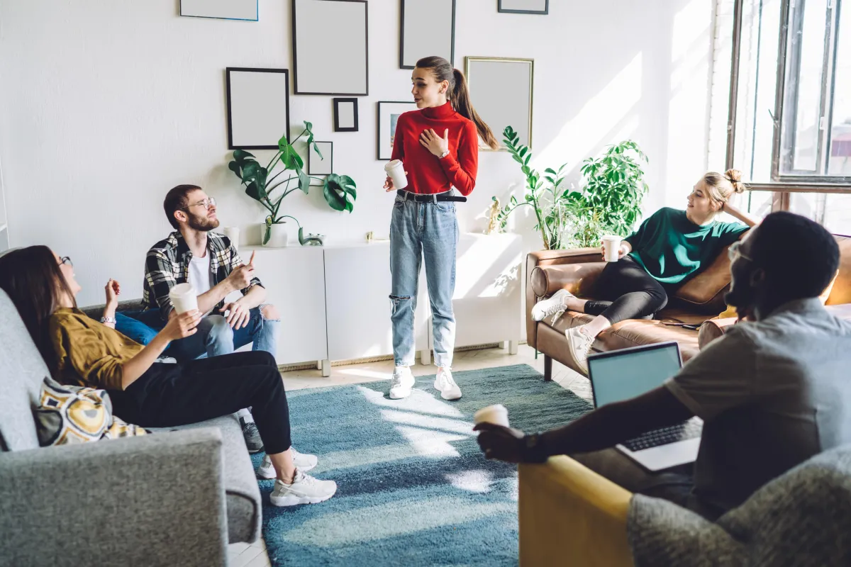 Young professionals having a casual team meeting in a modern, sunlit office lounge with coffee and laptops.