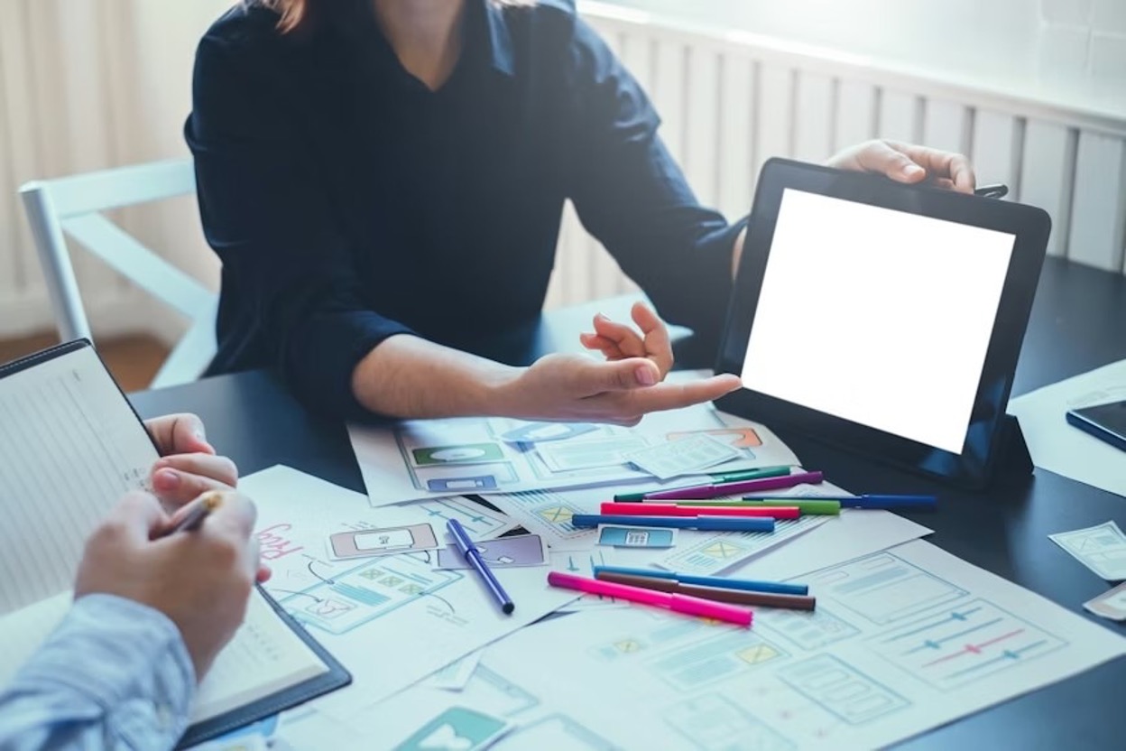 Two tech professionals are working on prototype development, one displaying a blank tablet screen and the other taking notes.