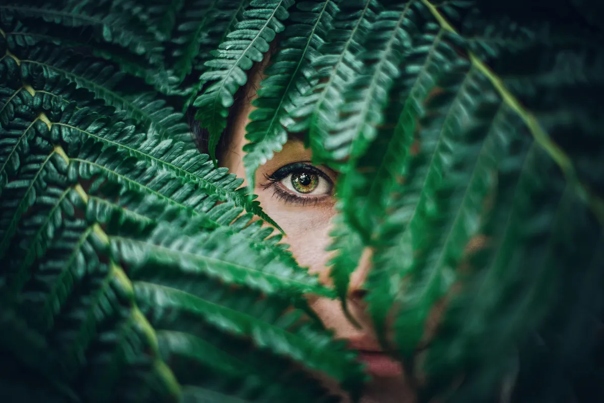 Close-up of a woman’s eye peeking through green fern leaves, symbolizing nervous system healing, emotional awakening, and the journey of self-regulation | Uhkare Mind Body Soul.